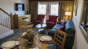 Quarto de hotel com área de jantar, sofá, televisão e varanda com cortinas.