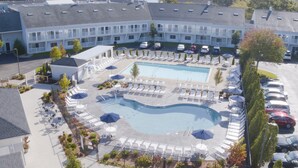 Um hotel com piscina cercada por espreguiçadeiras e guarda-sóis, estacionamento e um edifício com várias varandas.