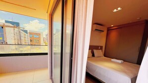 Ein Hotelzimmer mit Bett, Fenster mit Vorhängen, Blick auf Gebäude und einer Klimaanlage.