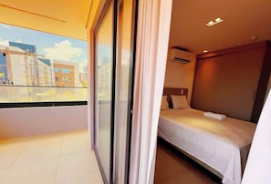 A hotel room with a bed, a window with curtains, a view of buildings, and an air conditioning unit. - Altamare - Miramar Hospedagens  (João Pessoa)