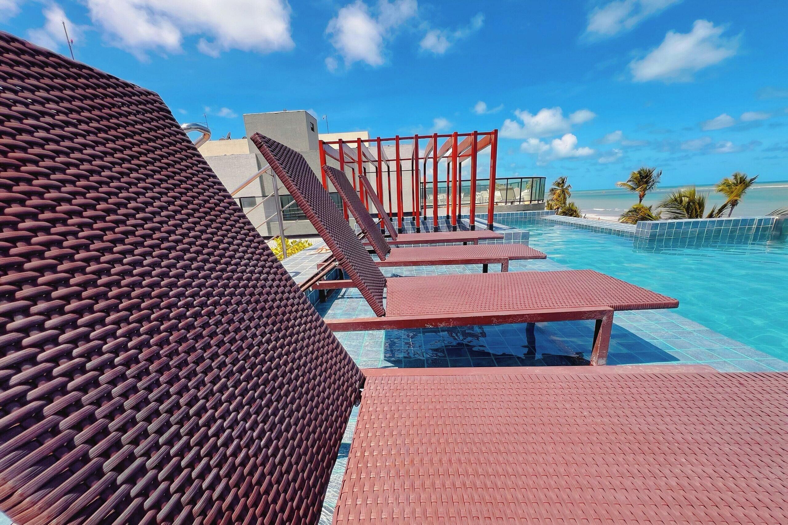 An infinity pool with red-tiled deck and lounge chairs, overlooking a beach.
