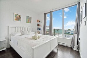 A white bedroom with a large window, a bed, a bedside table, and a radiator. - Modern 1-Bed Condo | Mountain Views | 10 mins > DT (Vancouver)