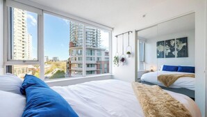 A modern bedroom with a large window, a bed with white bedding, a blue pillow, a fluffy rug, a world map on the wall, and a hanging plant. - Downtown Luxurious 2BD|Pool |Parking|Hot Tub|Sauna (Vancouver)