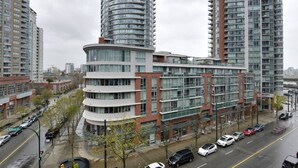 A modern high-rise building with multiple balconies, a curved corner design, and a street view with parked cars. - Downtown Luxurious 2BD|Pool |Parking|Hot Tub|Sauna (Vancouver)