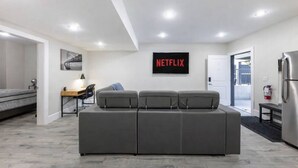 A modern, well-lit room with a grey sofa, a desk with a lamp, a wall-mounted TV displaying 'NETFLIX,' and a refrigerator. - Modern 2BR Retreat |Free Parking |Desk| Fast Wi-Fi (Surrey)