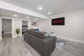 A modern living room with a grey sofa, a small side table, a plant, and a wall-mounted TV displaying 'NETFLIX'. - Modern 2BR Retreat |Free Parking |Desk| Fast Wi-Fi (Surrey)