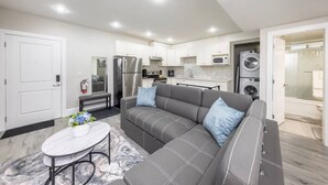 A modern apartment with a grey sofa, a small dining table, a kitchen with white cabinets, a built-in oven, and a washing machine. - Modern 2BR Retreat |Free Parking |Desk| Fast Wi-Fi (Surrey)
