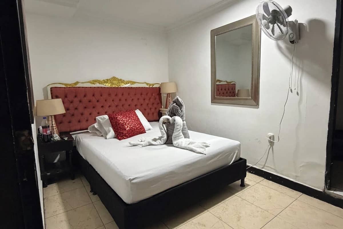 A hotel room with a bed, a nightstand, a lamp, a mirror, and a wall-mounted fan.