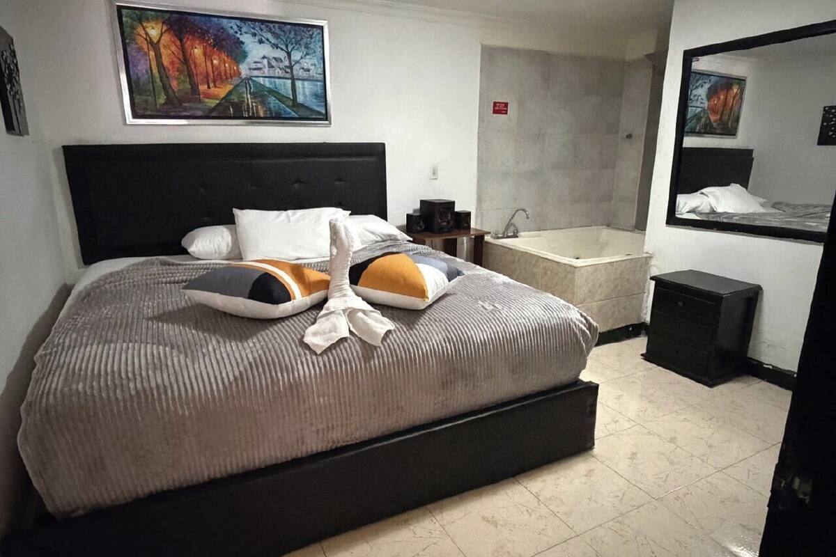 A bedroom with a large bed, a headboard, a painting, a bathtub, and a small table.