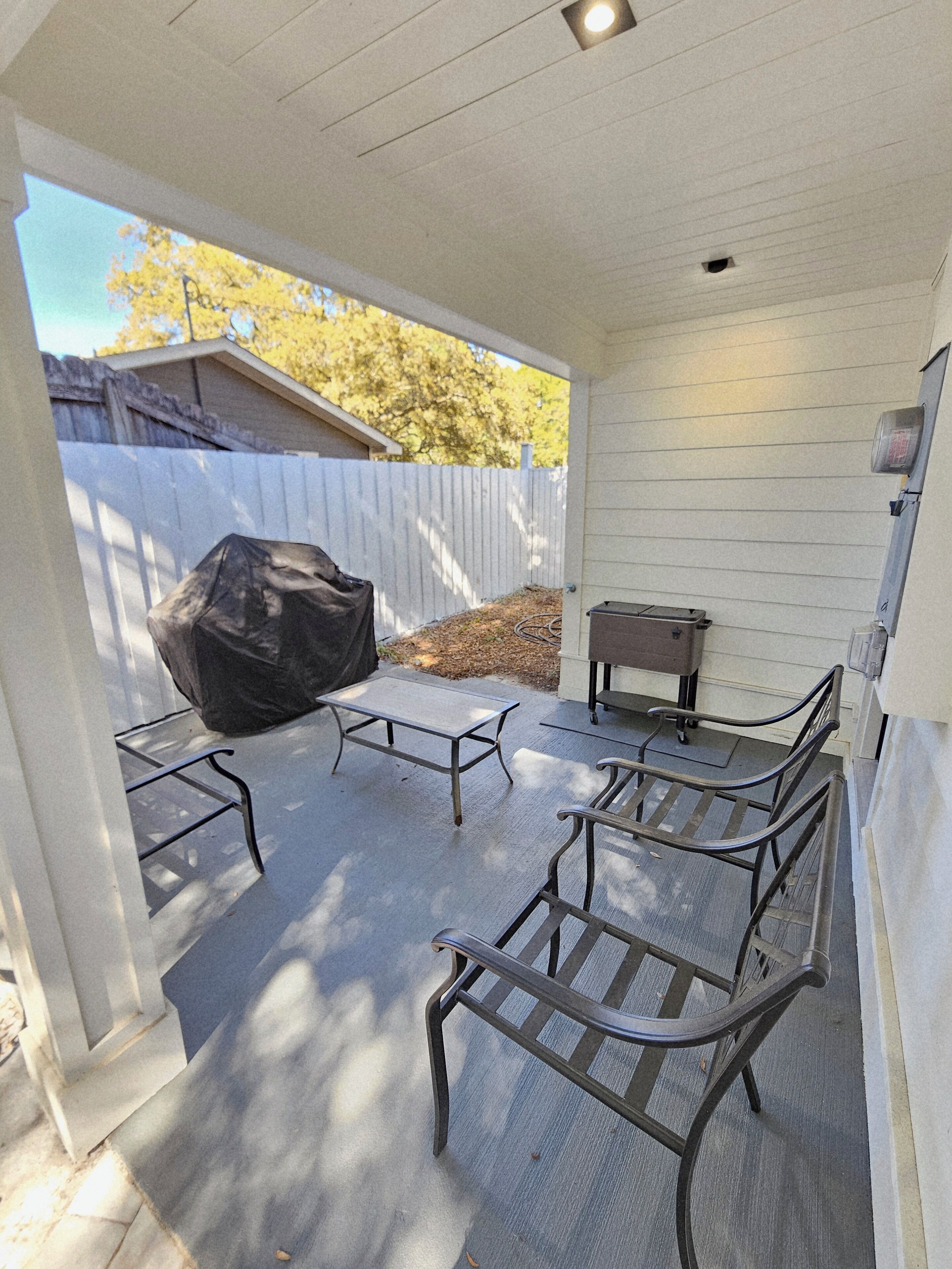 A covered patio with metal chairs and a table, a grill, and a white fence in the background.