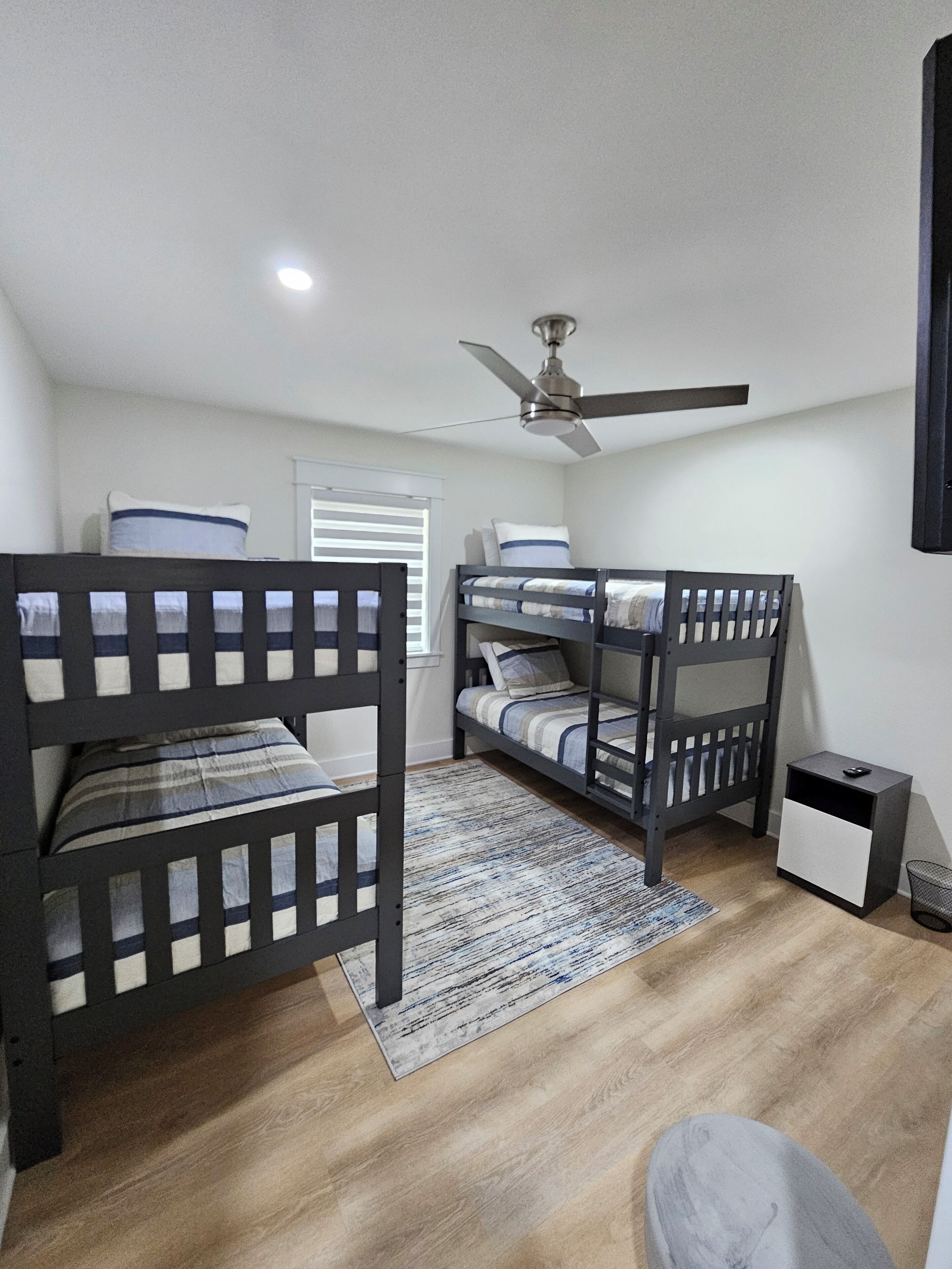 Ein Zimmer mit zwei Etagenbetten, einem Deckenventilator, einem kleinen Nachttisch und einem Fenster mit Jalousien.