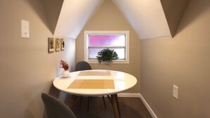 Een eetgedeelte met een ronde tafel, stoelen, een vaas met bloemen, een raam met een plant en een aan de muur hangend kunstwerk.