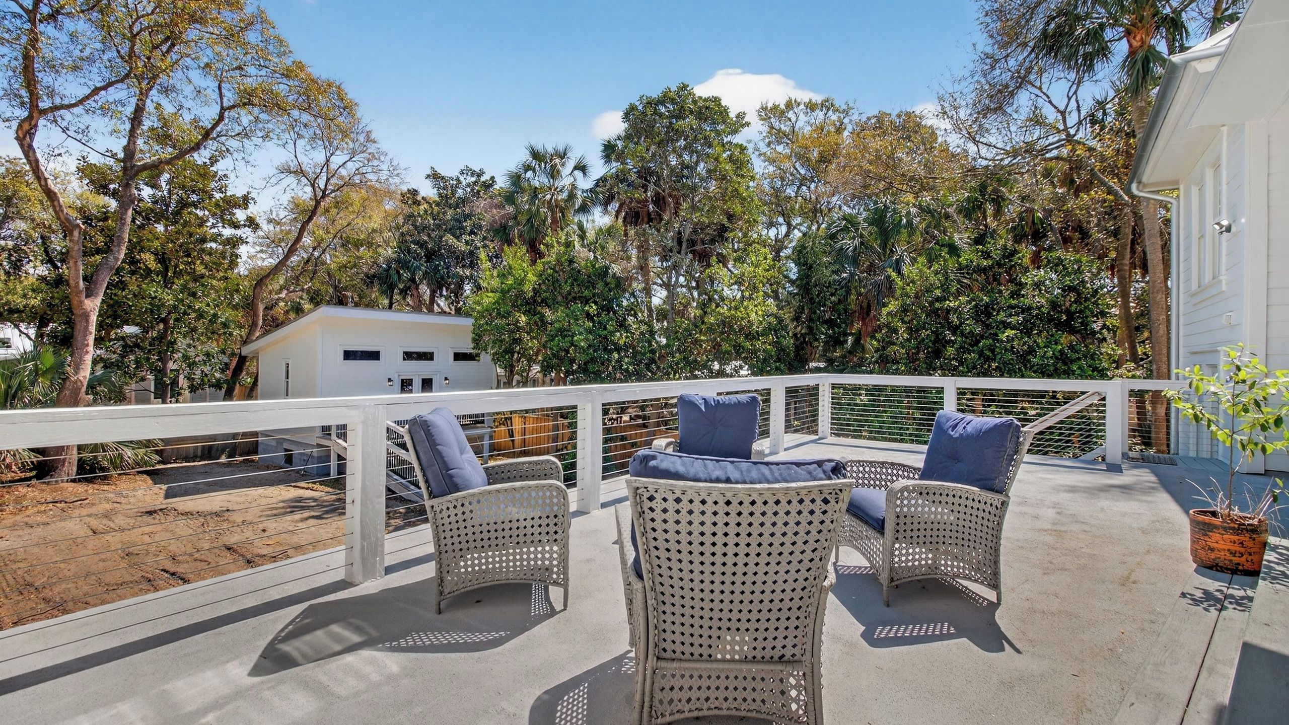 A covered outdoor seating area with wicker furniture and cushions, overlooking a garden with trees and a white building.
