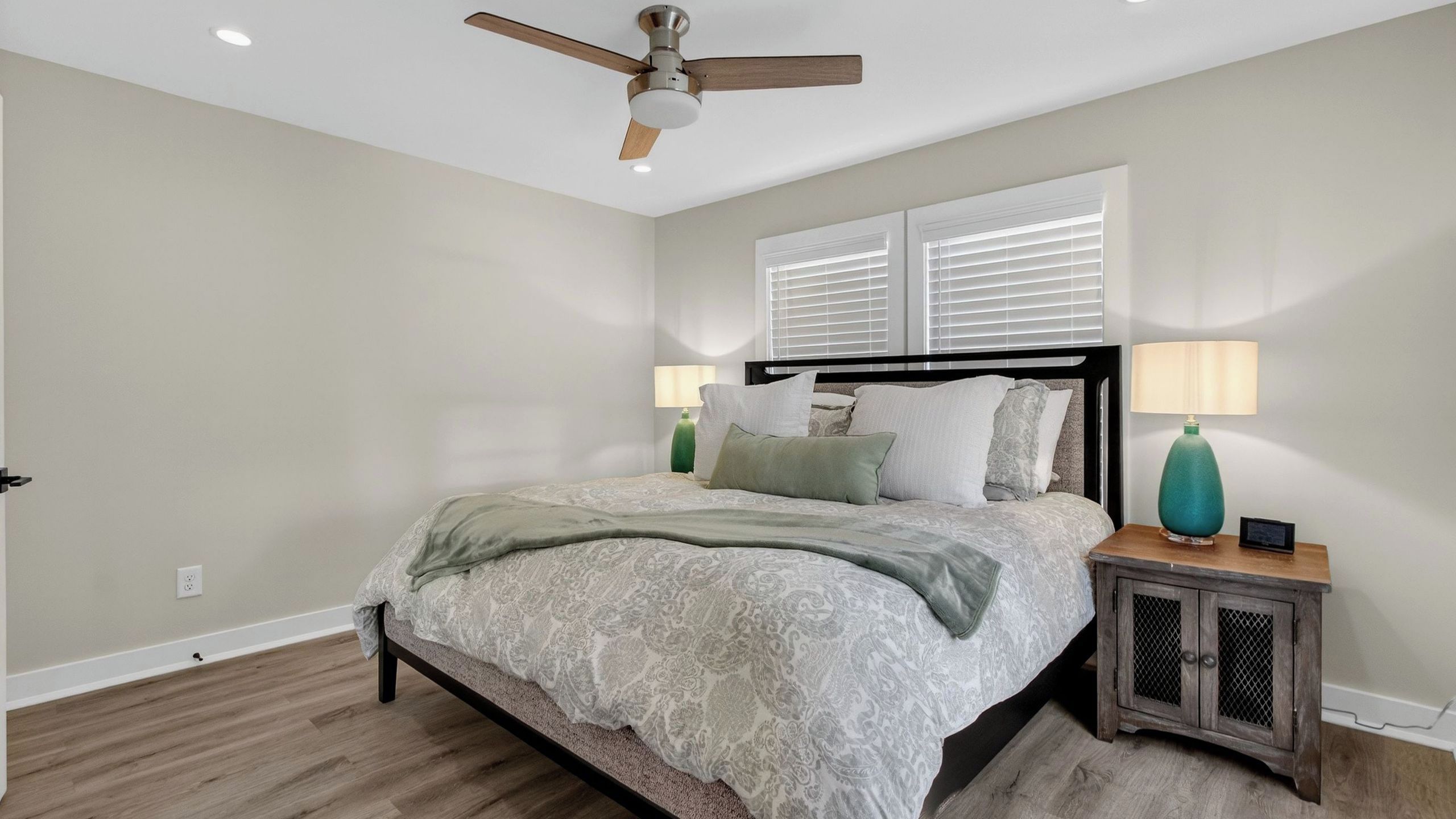 Une chambre à coucher comprenant un lit, une table de chevet, une lampe, un ventilateur de plafond et une fenêtre avec des stores.
