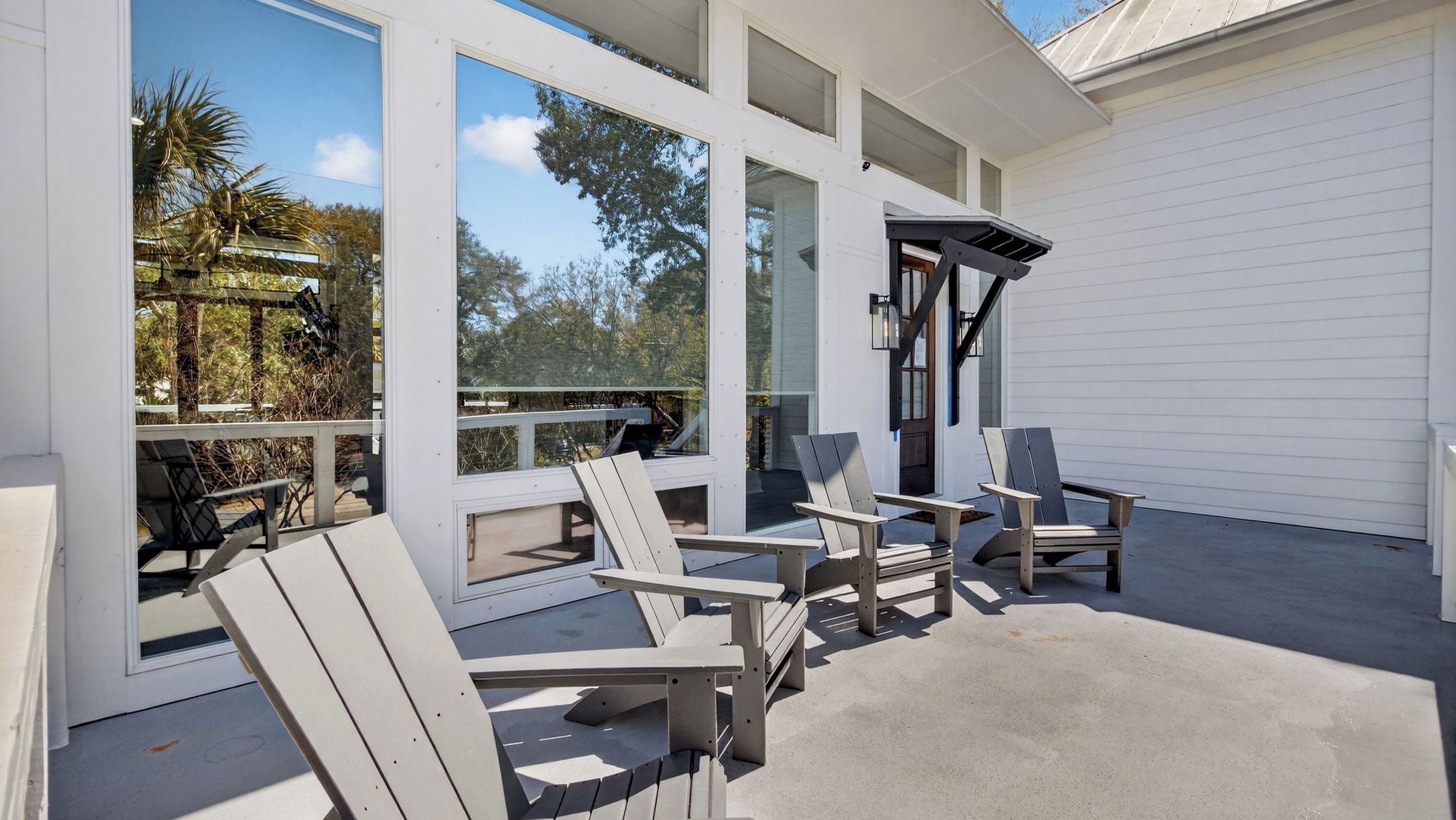 A covered outdoor area with Adirondack chairs and a table, large windows, and a view of trees.