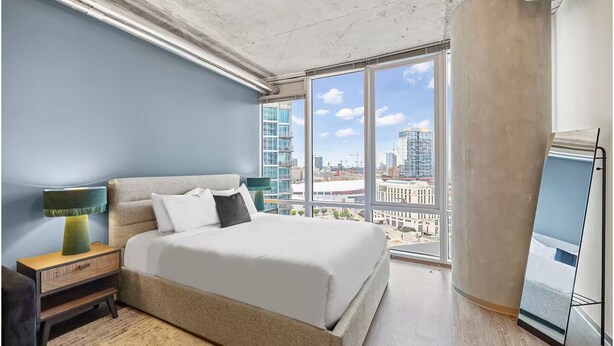 Ein modernes Hotelzimmer mit einem großen Bett, einem Nachttisch, einer Lampe und Blick auf die Stadt durch große Fenster.