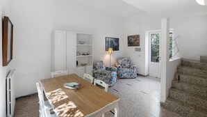 Ein Zimmer mit Esstisch, Stühlen, einem weißen Schrank, zwei blumigen Sesseln, einer Lampe und einer Treppe.