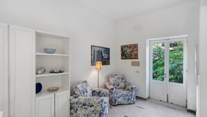 Ein Zimmer mit weißen Wänden, einer weißen Tür, zwei floralen Ohrensesseln, einer Stehlampe und einem Gemälde.