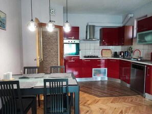 A modern kitchen with red cabinets, a stainless steel dishwasher, and a wooden dining table with chairs. - One Bedroom Apartment Sleeps 2 - Private Garden (Zadar)