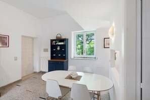 A room with a round table, white chairs, a bookshelf, a window with a view of trees, and a door. - Le Tore – Minucciola Apartment with Shared Terrace & Garden, Wi-Fi (Massa Lubrense)