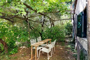 Ein Holztisch und weiße Stühle unter einem Baum in einem Garten mit einem Gebäude im Hintergrund.