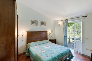 A bedroom with a bed, a wooden door, a nightstand, a wall-mounted light, and a window with curtains. - Le Tore – Palmento Room with Private Terrace, Shared Garden and Wi-Fi (Massa Lubrense)