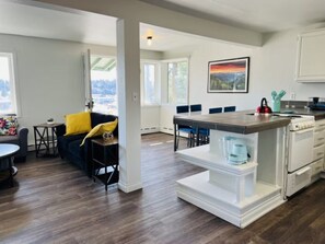 A modern kitchen with white cabinetry, a central island, and a dining area with blue chairs. - The Saltwater-OceanView,Downtown (Kodiak)
