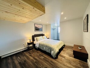 A bedroom with a wooden ceiling, a bed with a patterned bedspread, a bedside table with a lamp, and a small nightstand. - The Saltwater-OceanView,Downtown (Kodiak)