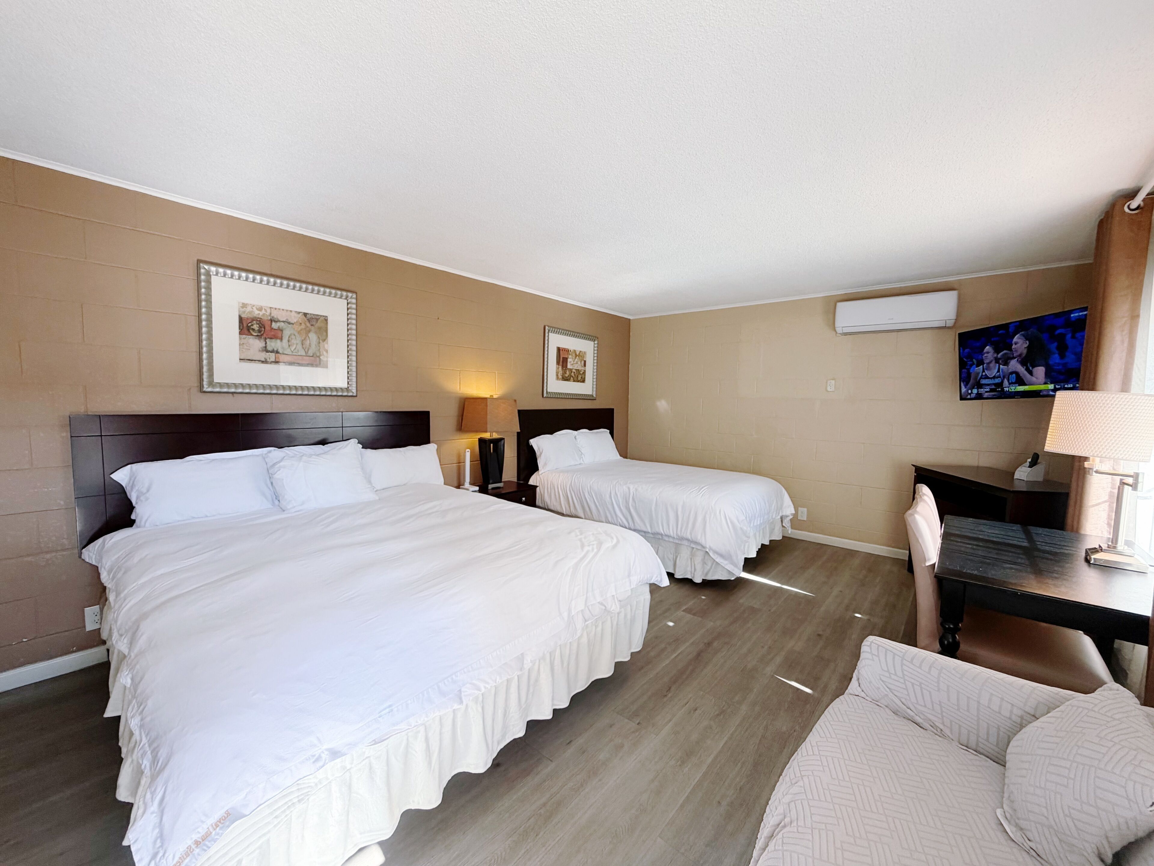 A hotel room with two beds, a desk, a chair, a TV, and framed artwork on the walls.