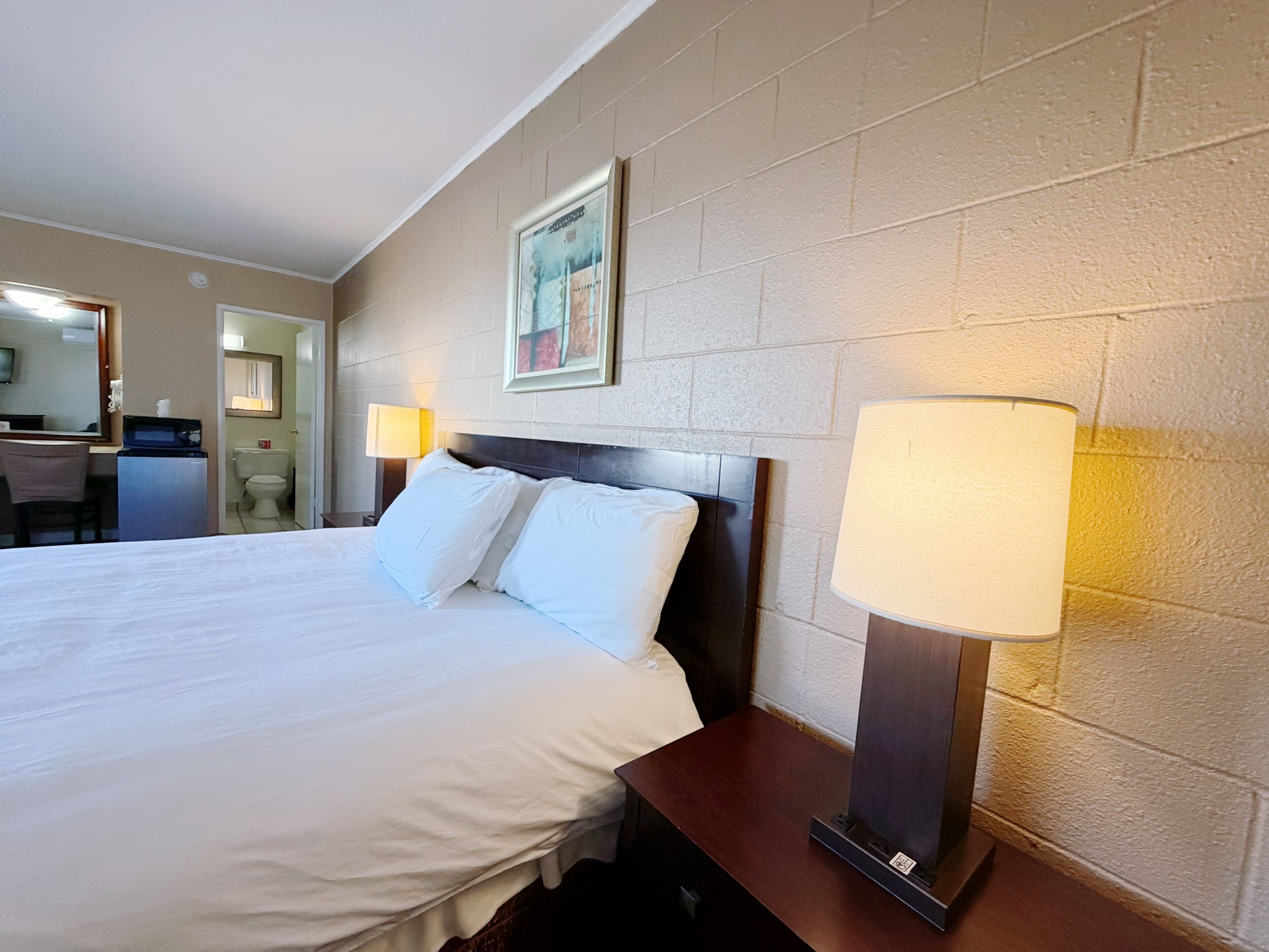 A hotel room with a large bed, bedside lamps, a framed picture on the wall, and a small desk with a chair.