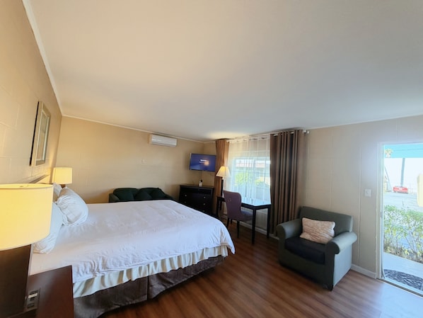 A hotel room with a bed, a desk, a chair, a television, and a view through a window. - Royal Inn & Suites (Hemet)