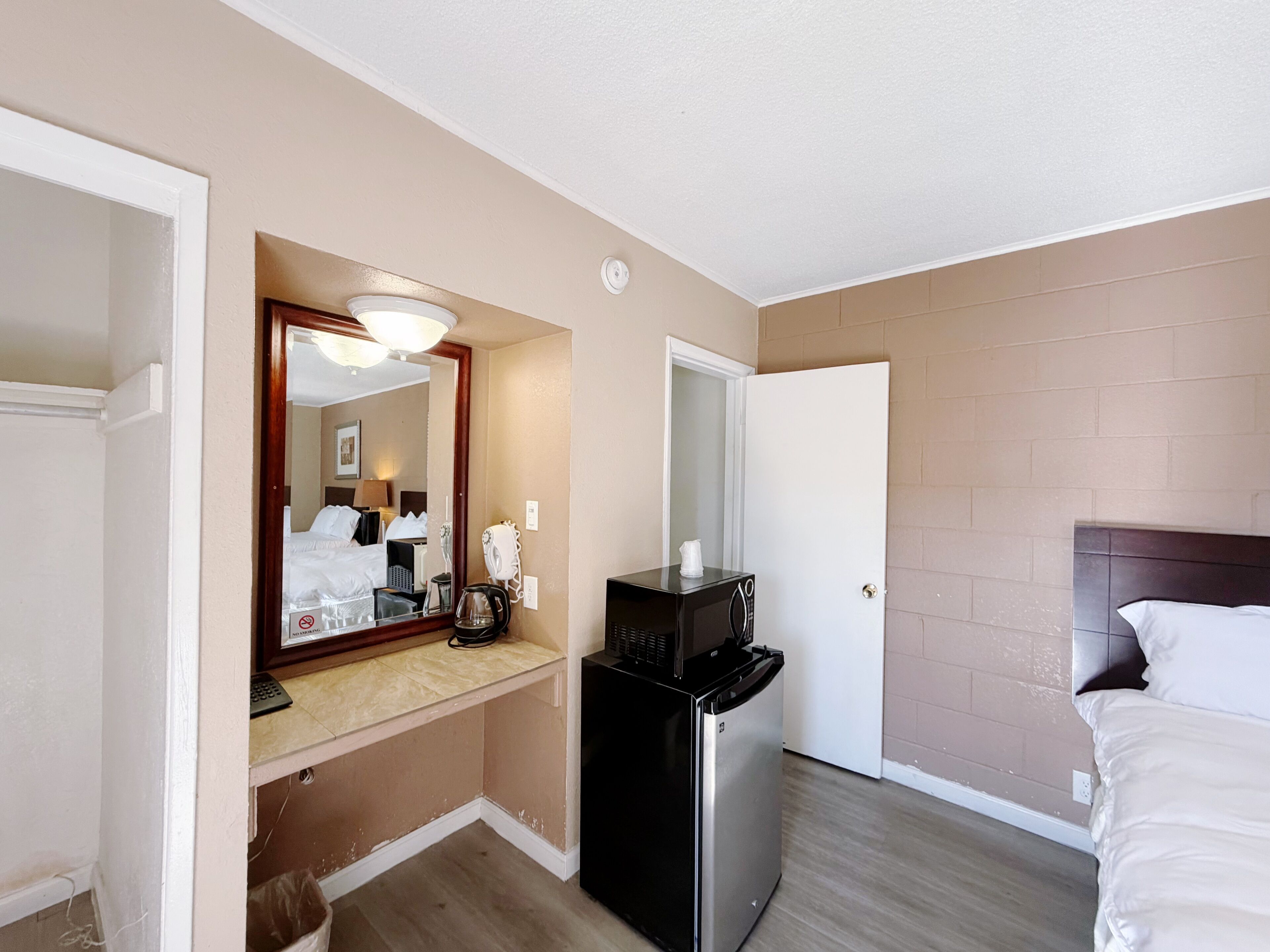 Quarto de hotel com cama, geladeira, micro-ondas, cafeteira, espelho e porta.