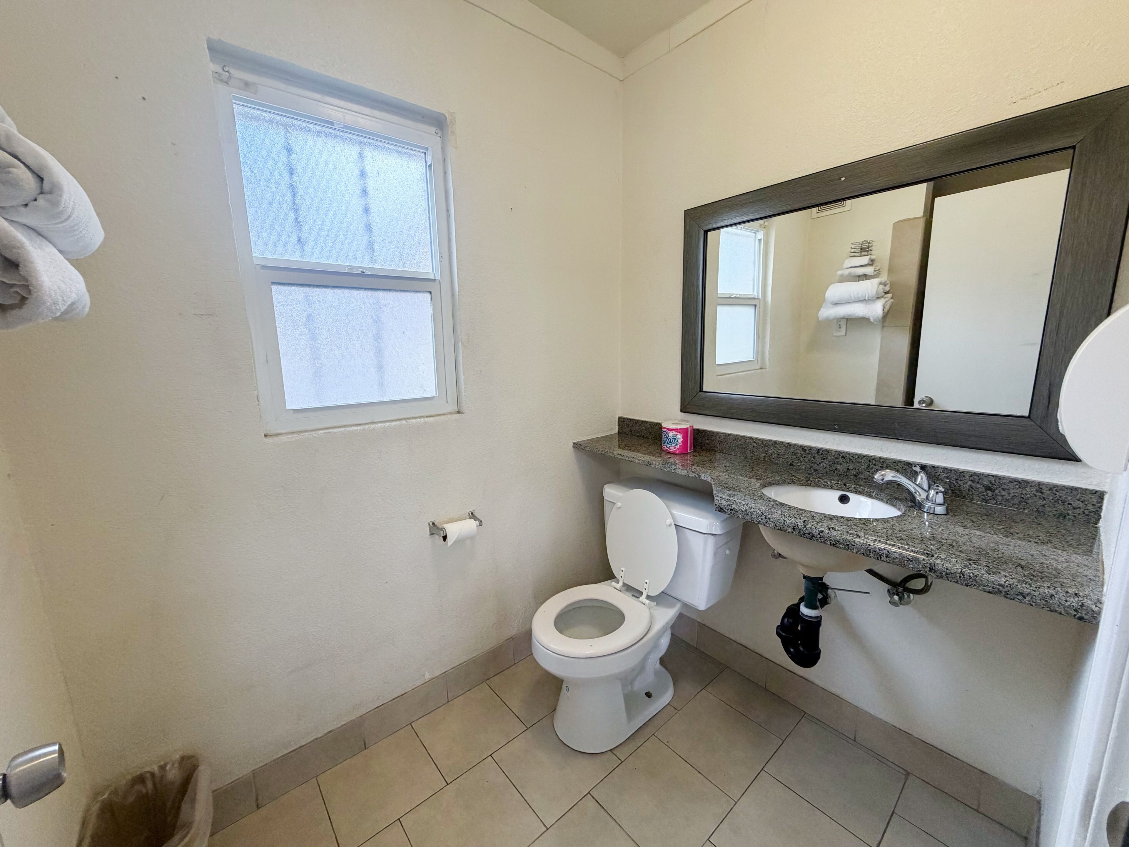 A bathroom with a toilet, sink, mirror, window, and a trash bin.