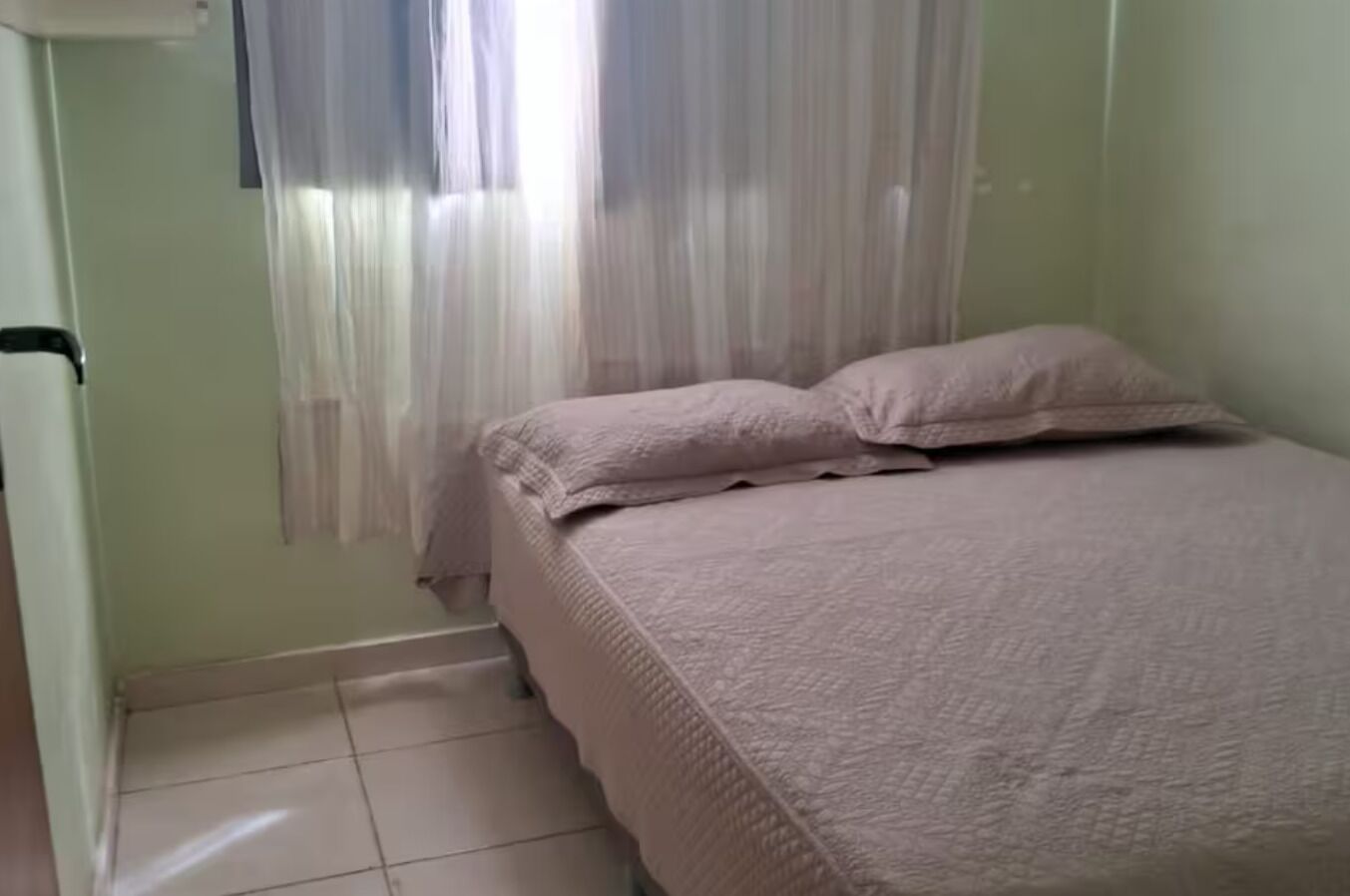 Una camera da letto con un letto, cuscini, una finestra con tende e un pavimento piastrellato.
