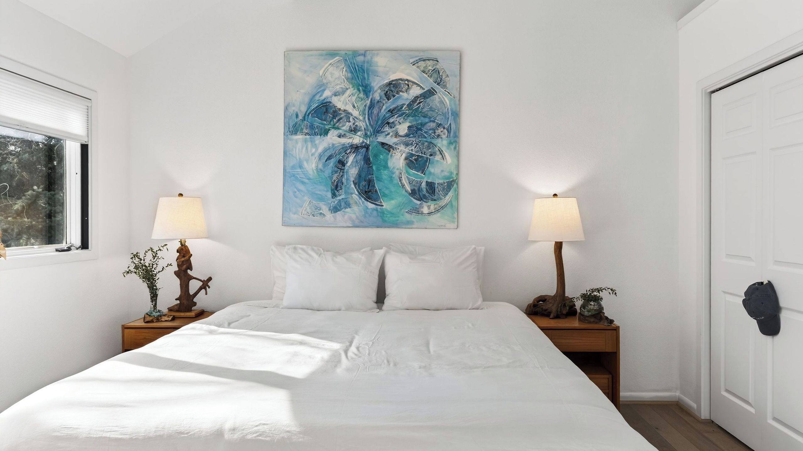 A bedroom with a bed, two bedside tables with lamps, a wall-mounted painting, and a window.
