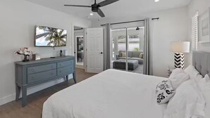 Et soveværelse med en seng, en kommode, en ventilator i loftet, et fjernsyn og en skydedør, der fører ud til en balkon.