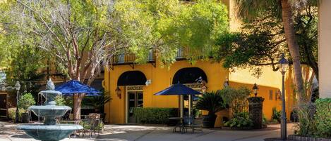 A courtyard with a fountain, outdoor seating, and a yellow building with arched windows.