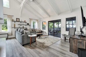 Un salon spacieux avec un plafond haut, un canapé, une table basse, un bureau avec un ordinateur et une vue à travers une porte vitrée.