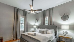 A bedroom with a bed, a ceiling fan, a bedside table with a lamp, and a window with curtains.
