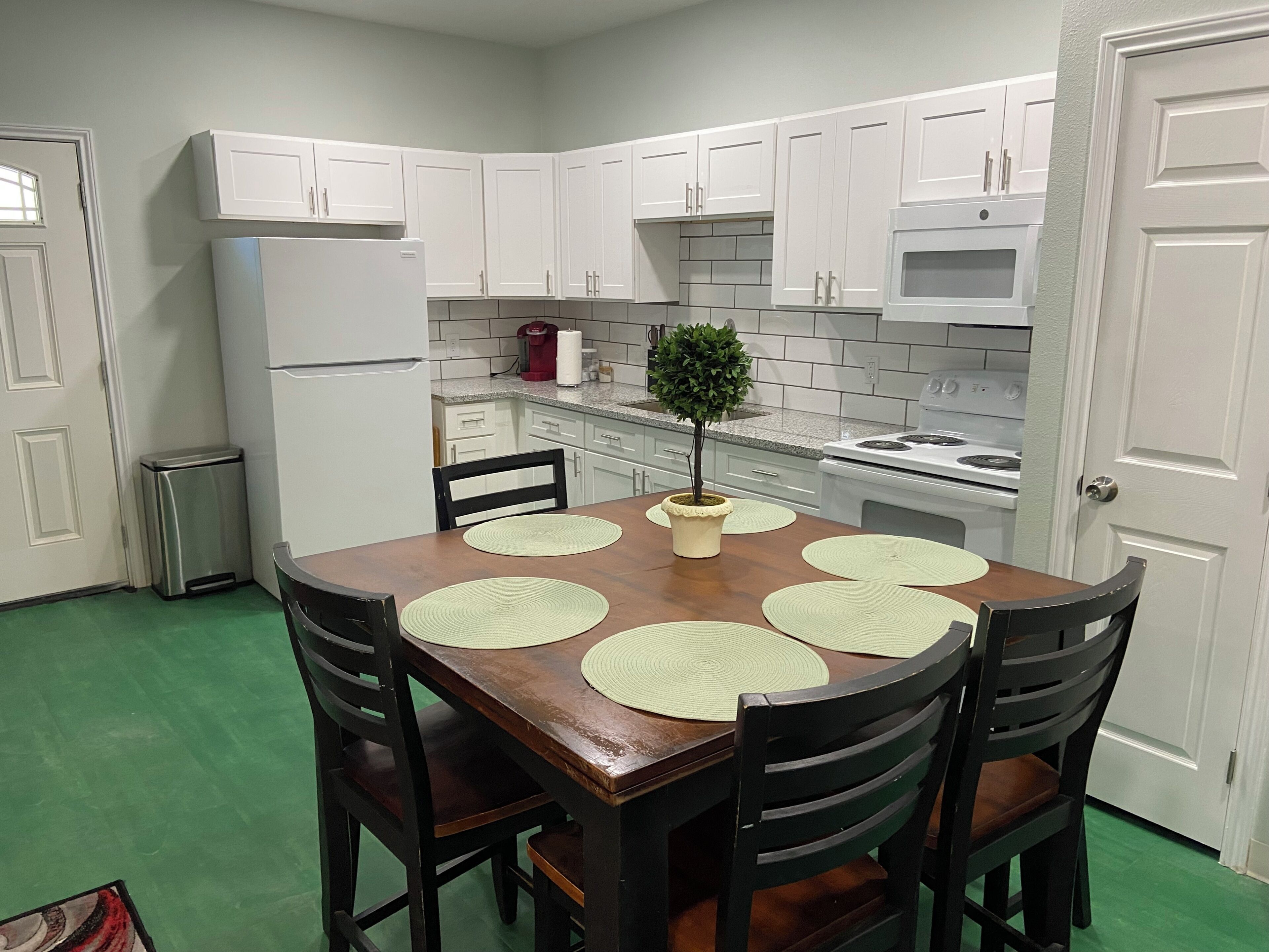 A compact kitchen with white cabinets, a refrigerator, a microwave, and a stove. There is a dining table with four chairs and a small potted plant.