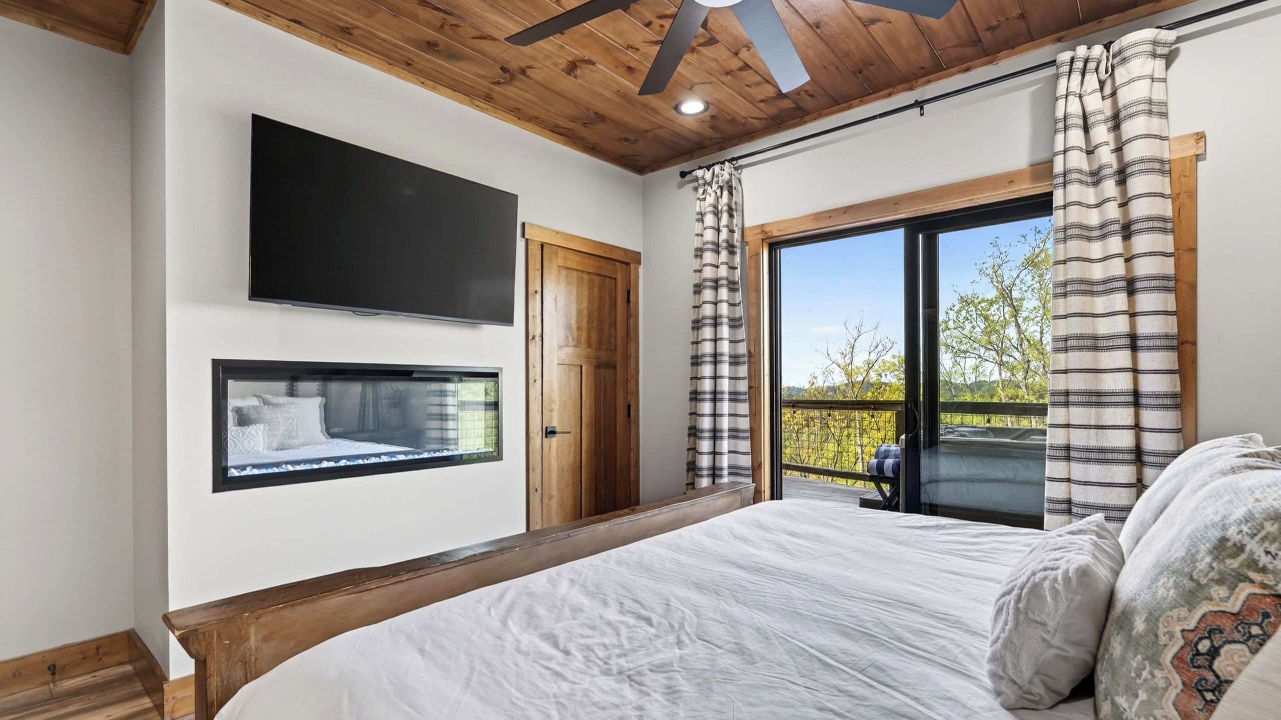 A bedroom with a bed, a mounted TV, a window with curtains, and a balcony with a view.