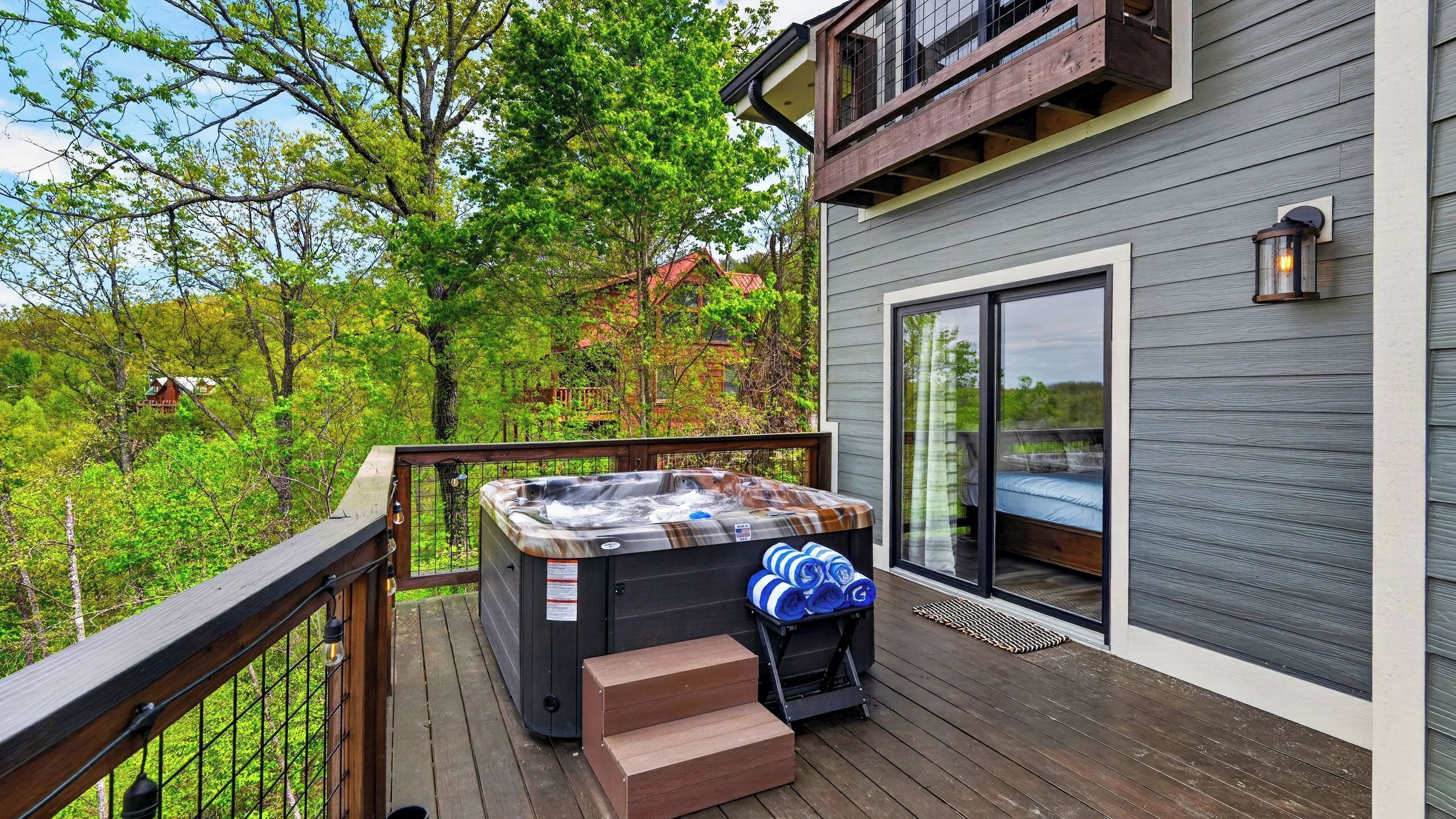 Un bain à remous sur une terrasse en bois, accessible par des marches menant à une porte vitrée, le tout entouré d’arbres.