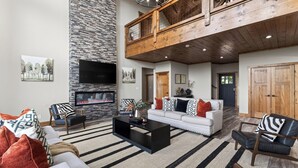 A spacious living room with a stone wall, a fireplace, a black TV, and a high ceiling with exposed beams. - Priddy Secluded by Avantstay Hot Tub + Expansive Deck + Game Room + Home Theater (Sevierville)