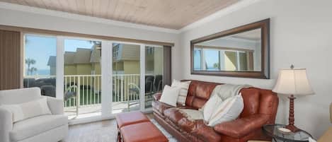 A spacious living room with a brown leather sofa, a white armchair, and a bench. There is a large window with a view of a balcony and buildings.