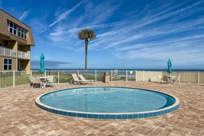 Una piscina amb gandules i ombrel·les, envoltada per una tanca, amb vista a l'oceà i un edifici al fons.