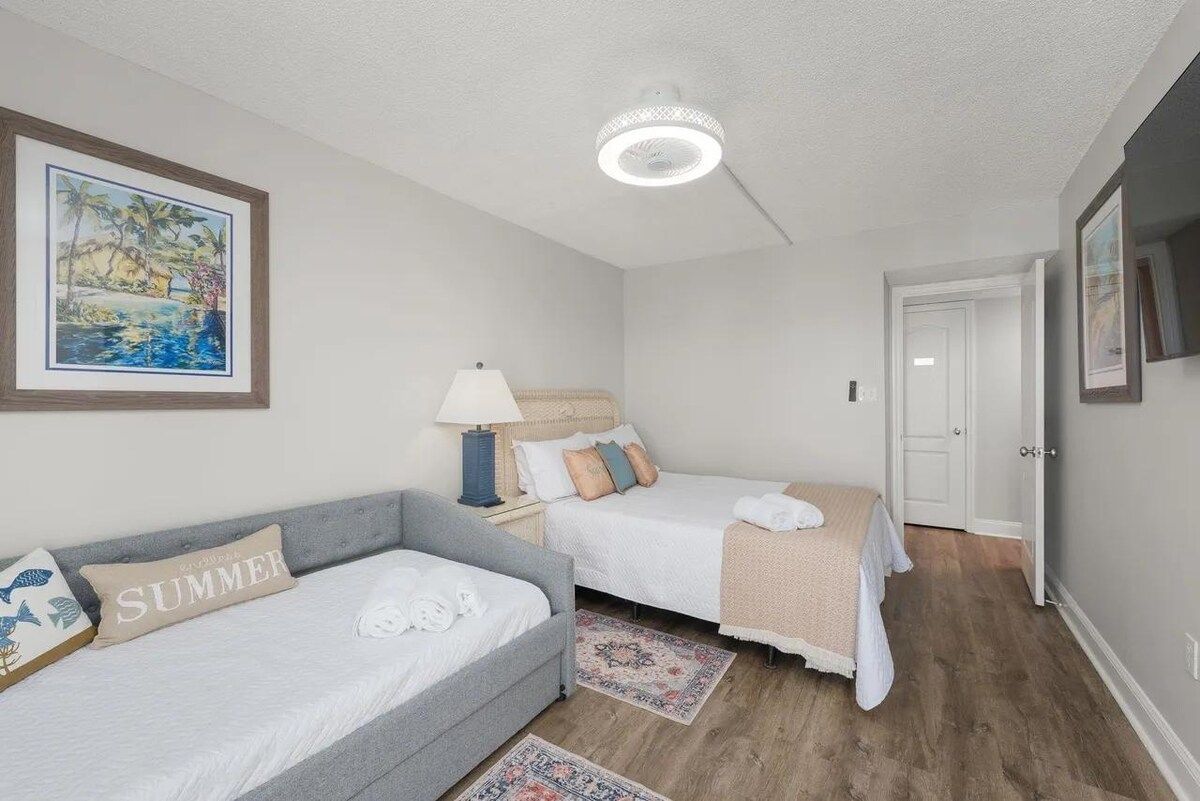 A bedroom with two beds, a nightstand, a lamp, a framed picture, and a door.
