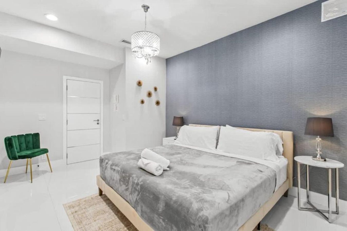 A modern bedroom with a bed, bedside lamps, a green chair, and a chandelier.