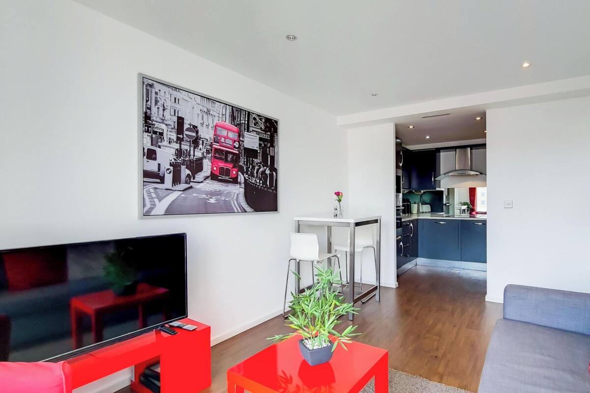 Una sala de estar moderna con un televisor de pantalla plana, una mesa de centro roja con una planta, un sofá gris con cojines rojos y blancos, y una cocina al fondo.