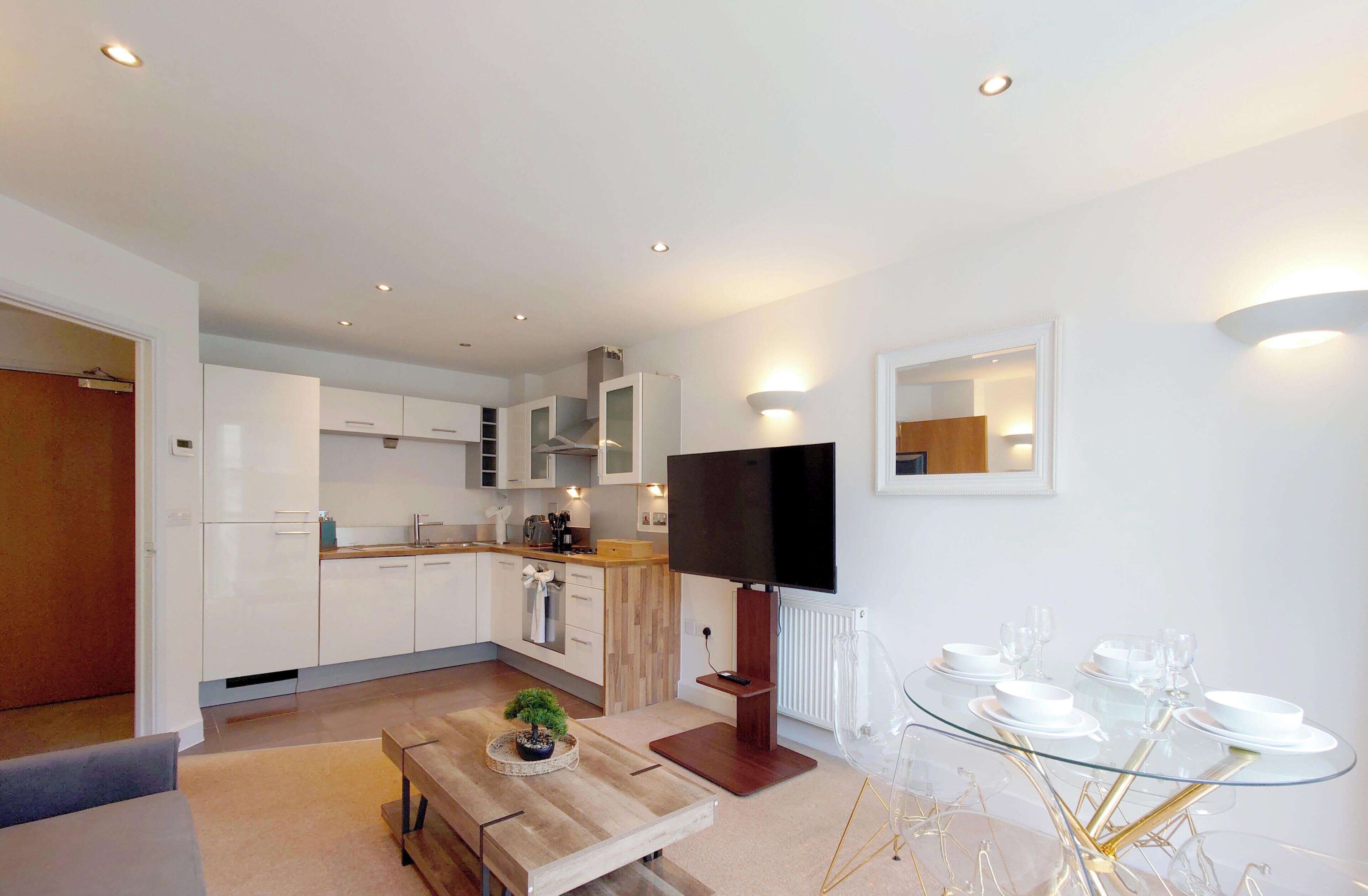 A modern kitchen with white cabinets, a dining area with a glass table, and a flat-screen TV mounted on the wall.