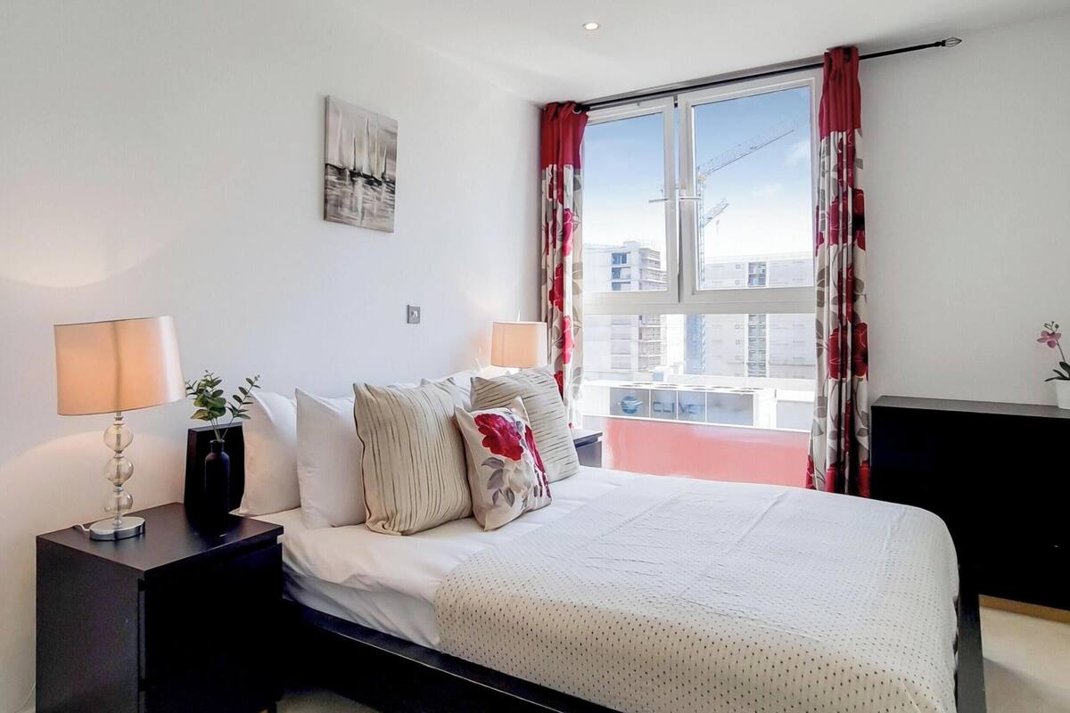 Un dormitorio con cama, lámparas de noche, una ventana con cortinas, una planta pequeña y una obra de arte en la pared.